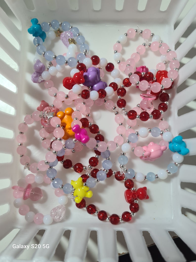 K&ouml;nnte beinhalten: Eine Sammlung bunter Perlenarmb&auml;nder mit verschiedenen Anh&auml;ngern, darunter B&auml;ren, Herzen und Sterne. Die Armb&auml;nder sind aus rosa, roten, blauen und wei&szlig;en Perlen gefertigt.