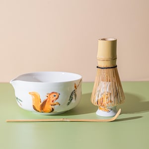 Op de afbeelding: Een witte keramische matcha kom met een eekhoorn illustratie, een bamboe klopper en een bamboe lepel. De eekhoorn is oranje en bruin. De klopper is gemaakt van bamboe en heeft een zwart touwtje.