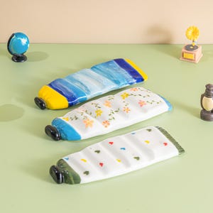 Puede incluir: Tres fuentes de cerámica con forma de tubo de pintura. Una es azul y amarilla, otra blanca con diseños florales y la tercera blanca con patrones coloridos de corazones y puntos. Las fuentes están sobre una superficie verde claro.