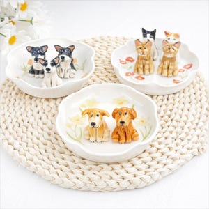 K&ouml;nnte beinhalten: Drei dekorative Keramikschalen mit gewelltem Rand, jeweils mit Hunde- und Katzenfiguren. Die Schalen sind wei&szlig; mit floralen Akzenten. Die Hunde und Katzen sind in verschiedenen Farben und Posen dargestellt.
