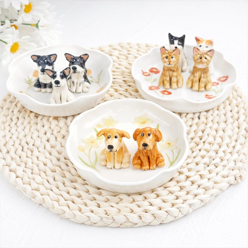 K&ouml;nnte beinhalten: Drei dekorative Keramikschalen mit gewelltem Rand, jeweils mit Hunde- und Katzenfiguren. Die Schalen sind wei&szlig; mit floralen Akzenten. Die Hunde und Katzen sind in verschiedenen Farben und Posen dargestellt.