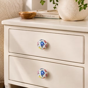 May include: Two round white ceramic drawer knobs featuring a hand-painted floral pattern with blue, red, and yellow petals and green leaves. The knobs are installed on a cream-colored wooden nightstand with two drawers, providing a decorative accent.