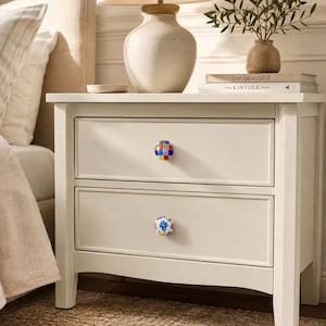 May include: A white wooden nightstand with two drawers, each featuring a unique decorative knob. The top drawer has a multicolored checkered knob, while the bottom drawer has a white knob with a blue and red floral pattern design.