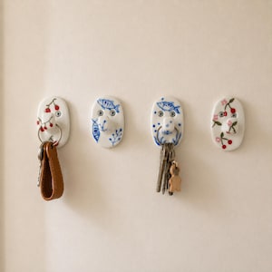 May include: Four oval ceramic wall hooks mounted with screws. The first features red berries, the second blue fish and foliage, the third blue fish and dots, and the fourth pink flowers and red cherries. Two hooks hold metal key rings with keys.