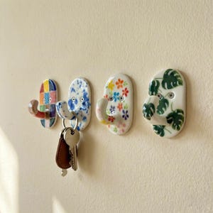 Puede incluir: Cuatro ganchos de pared de cerámica con diseños decorativos. Uno tiene un patrón geométrico colorido, otro tiene salpicaduras azules y blancas, un tercero tiene patrones florales y el último tiene patrones de hojas verdes. Las llaves cuelgan de un gancho.