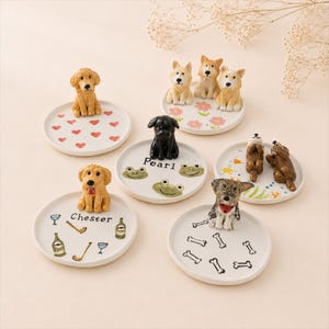 Puede incluir: Una colección de figuras de perros de cerámica sobre pequeños platos redondos. Cada plato presenta un diseño único, que incluye corazones, flores, caras de rana y huesos de perro. Algunos platos tienen nombres como "Chester" y "Pearl".