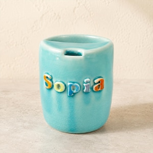 Puede incluir: Una taza de cerámica azul claro con un acabado de esmalte craquelado brillante. El nombre "Sopia" está grabado en el frente en letras multicolores en relieve con tonos naranja, amarillo y azul. La taza tiene una forma cilíndrica redondeada.