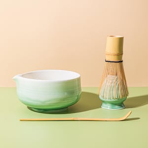 Puede incluir: Un juego de té matcha con un cuenco de cerámica verde claro con pico, un batidor de bambú y una cuchara de bambú. El cuenco tiene un interior blanco y un degradado de tonos verdes. El batidor es de bambú.