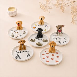Puede incluir: Conjunto de platos decorativos de cerámica blanca, cada uno con una figura de perro diferente. Los platos presentan varios diseños, incluyendo corazones, huesos y caras de rana. Algunos platos tienen nombres como "Chester" y "Pearl".