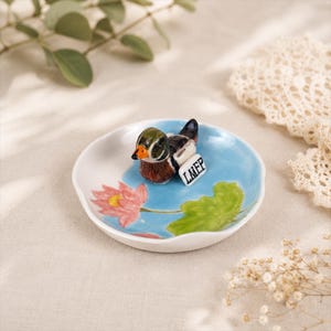 Puede incluir: Un peque&ntilde;o plato de cer&aacute;mica blanco con un dise&ntilde;o pintado a mano que presenta una figura de pato. El pato es marr&oacute;n, verde y negro con un pico naranja. El plato tiene un fondo azul con una flor de loto rosa y una hoja verde.