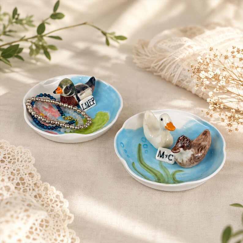 Puede incluir: Dos cuencos de cer&aacute;mica para baratijas con figuras de patos. Un cuenco tiene un dise&ntilde;o azul y blanco con un pato real y una pulsera. El otro cuenco tiene un pato blanco y un pato marr&oacute;n con las iniciales "M+C".