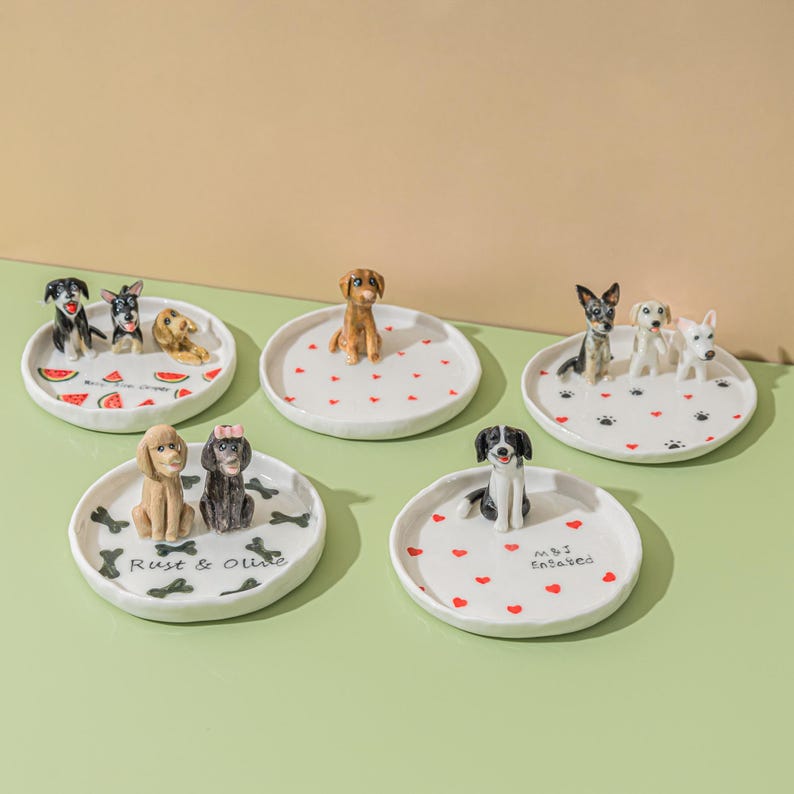 May include: A collection of white ceramic dishes, each adorned with a different dog figurine. Some dishes feature red hearts, watermelon slices, or bone designs. The text "Rust & Olive" and "M&J Endaved" are visible on two of the dishes.