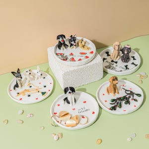 Op de afbeelding: Een verzameling witte keramische ringgerechten, elk versierd met een ander hondenbeeldje. Sommige gerechten hebben pootafdrukken, harten en watermeloenplakjes. Eén gerecht heeft de tekst "M&I Entwined". Goudkleurige sieraden liggen op één gerecht.