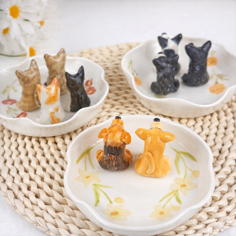 K&ouml;nnte beinhalten: Eine Sammlung von Keramik-Hundefiguren in verschiedenen Farben, darunter Braun, Schwarz und Wei&szlig;, wird in dekorativen Schalen mit gewelltem Rand pr&auml;sentiert. Die wei&szlig;en Schalen sind mit Blumenmustern versehen und stehen auf einer gewebten Matte.