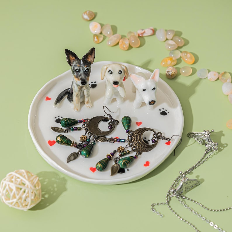 May include: A white ceramic plate with three dog figurines and two pairs of earrings. The earrings have a crescent moon design with green and gold accents. The plate is decorated with paw prints and red hearts.