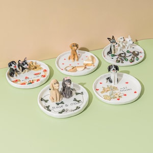 May include: A collection of white ceramic trinket dishes, each adorned with a different breed of dog figurine. Some dishes feature additional decorative elements like hearts, bones, and the text "Rust & Olive" and "M.B.I. Entwined".