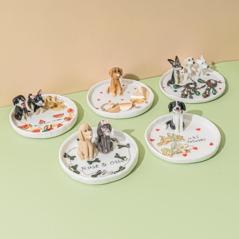 May include: A collection of white ceramic trinket dishes, each adorned with a different breed of dog figurine. Some dishes feature additional decorative elements like hearts, bones, and the text "Rust & Olive" and "M.B.I. Entwined".