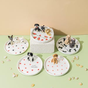 May include: A collection of white ceramic trinket dishes, each adorned with a different dog figurine. Some dishes feature paw prints, hearts, or watermelon slices. One dish has the text "M&J Entwined", and another reads "Rust & Olive".