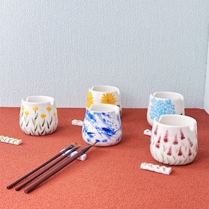 Puede incluir: Conjunto de botes de pintura de cerámica con varios diseños florales y abstractos. Los botes blancos presentan detalles en amarillo, azul y rojo. También se ven pinceles y pequeños soportes de cerámica para pinceles, lo que sugiere un tema artístico o artesanal.