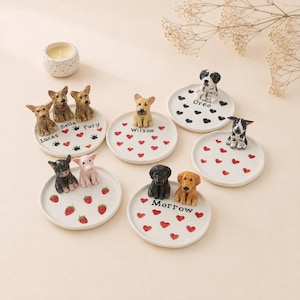 Puede incluir: Una colección de pequeños platos de cerámica blanca, cada uno adornado con una figura de perro en miniatura. Algunos platos presentan corazones rojos, huellas de patas negras o fresas. Nombres como "Lucas", "Bella" y "Oreo" están escritos en los platos.