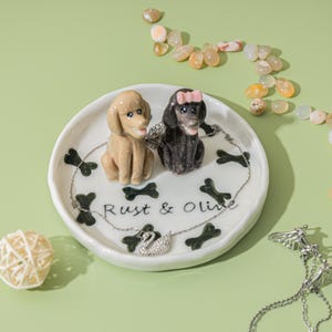 May include: A white ceramic trinket dish with a light brown poodle figurine and a black poodle figurine with a pink bow. The dish has black bone accents and the text "Rust & Olive". A silver swan necklace is also present.