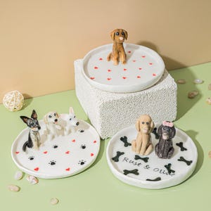 May include: White ceramic dog-themed trinket dishes. One dish features a brown dog figurine, while others have multiple dog figurines. The dishes are decorated with paw prints, hearts, and bone designs. One dish has the text "Rust & Olive".