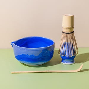 May include: A blue ceramic matcha bowl with a spout, a bamboo whisk, and a bamboo scoop. The bowl has a wave-like design in shades of blue. The whisk has a bamboo handle and bristles. The scoop is light brown.