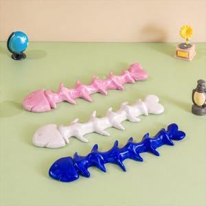 May include: Three decorative fish bone sculptures in pink, white, and blue. Each sculpture has a unique design, with a head and tail shape. The sculptures are placed on a light green surface.