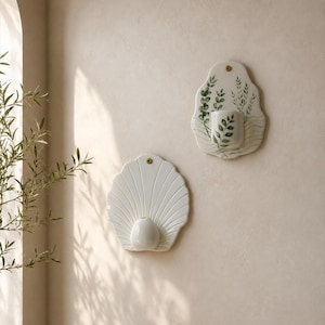 Op de afbeelding: Twee witte keramische wandplantenbakken. De ene heeft de vorm van een geschulpte schelp met een ronde houder. De andere heeft een groen botanisch bladpatroon met een cilindrische houder, beide bevestigd met messingkleurige schroeven.