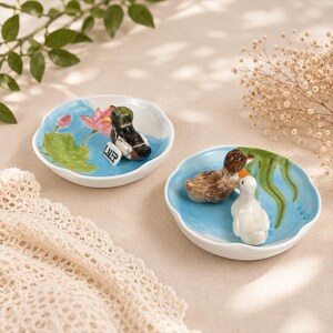 Puede incluir: Dos peque&ntilde;os platos de cer&aacute;mica blanca con borde ondulado. Uno presenta una figura de pato y una figura de patito blanco, con detalles de agua azul y plantas verdes. El otro plato tiene una figura de pato y un dise&ntilde;o de flor de loto.