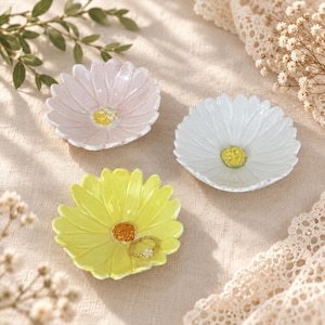 Puede incluir: Tres platos de cerámica para joyas con forma de margarita en rosa pastel, blanco y amarillo brillante. Cada plato presenta un centro texturizado y elevado en un color contrastante. Un pequeño anillo dorado con un detalle floral descansa en el plato amarillo.