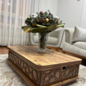 May include: A wooden coffee table with a decorative carved design around the edge. The table has a light brown wood finish and is sitting on a patterned rug.