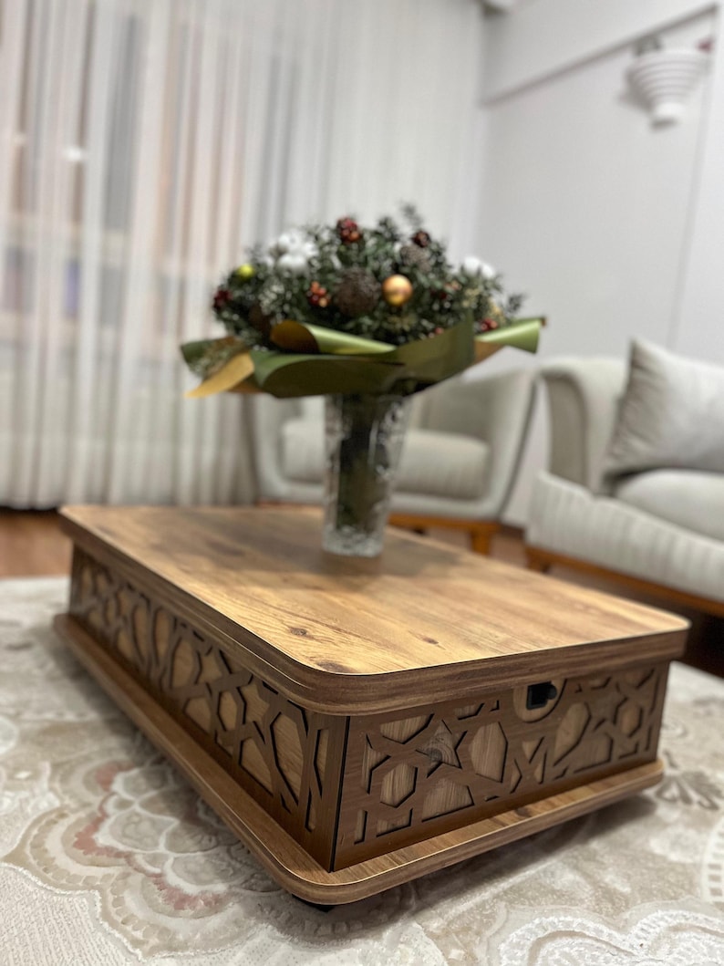 May include: A wooden coffee table with a decorative carved design around the edge. The table has a light brown wood finish and is sitting on a patterned rug.