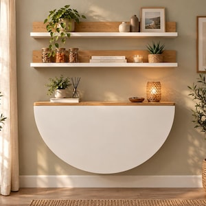 May include: A wall-mounted, semi-circular drop-leaf table featuring a white curved front panel and a natural wood-toned top surface. The table is positioned below two horizontal white shelves with wood backings, which hold various decorative items.