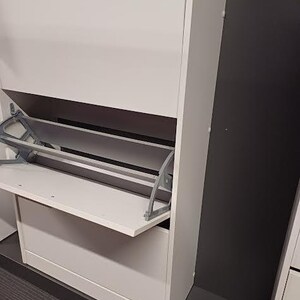 Op de afbeelding: Een witte schoenenkast met een scharnierende deur en twee planken. De deur staat open en laat een plank zien met een metalen steun.