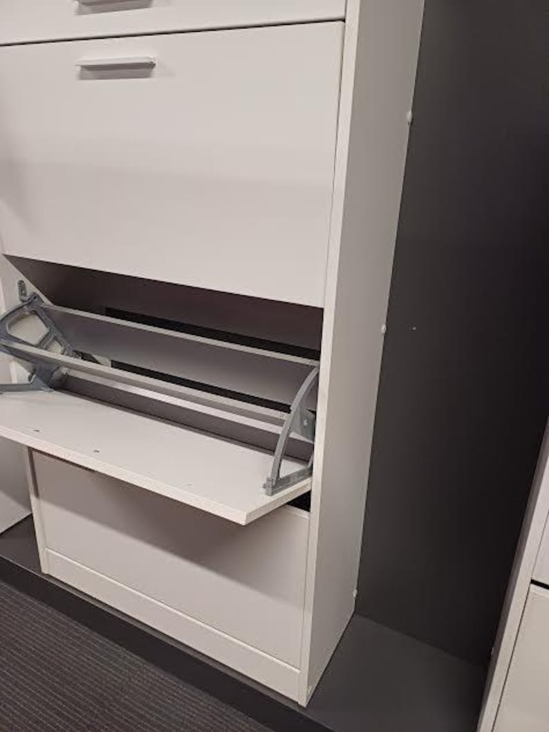 Op de afbeelding: Een witte schoenenkast met een scharnierende deur en twee planken. De deur staat open en laat een plank zien met een metalen steun.