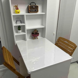 May include: White folding table with a white shelf unit above it. The shelf unit has three shelves and is attached to the wall. There are two wooden chairs in front of the table. The table has a clear plastic cover on top.