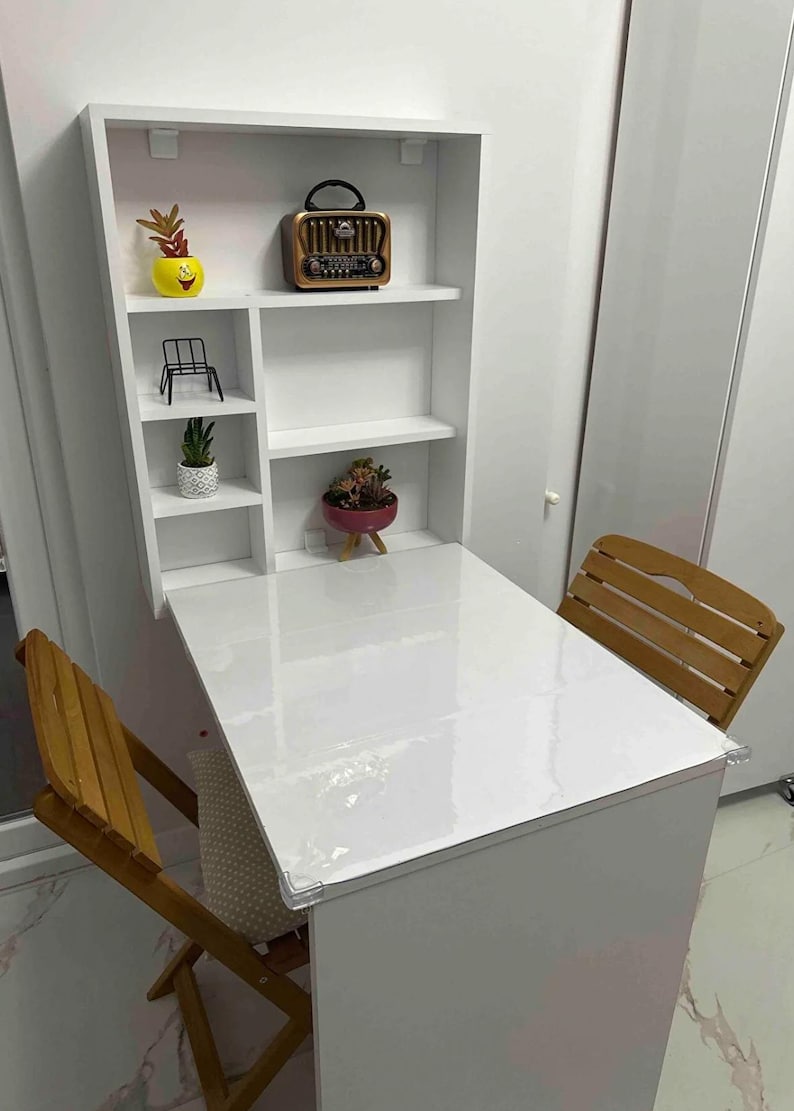 May include: White folding table with a white shelf unit above it. The shelf unit has three shelves and is attached to the wall. There are two wooden chairs in front of the table. The table has a clear plastic cover on top.