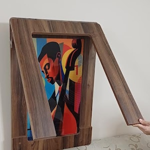 K&ouml;nnte beinhalten: Ein Holzrahmen mit einer klappbaren Frontplatte, der einen farbenfrohen abstrakten Druck einer Person zeigt, die ein Musikinstrument spielt. Der Druck zeigt eine Person mit dunkler Hautfarbe, die ein blaues Hemd tr&auml;gt und ein gro&szlig;es, rot, gelb und schwarzes Instrument spielt. Der Rahmen ist braun und hat eine nat&uuml;rliche Holzmaserung.