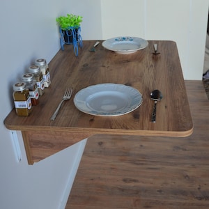 May include: A wall-mounted, wooden table with a light brown finish. Two white plates, silverware, and spice jars are arranged on the table. A small blue bicycle planter and a blue wall planter are also visible.