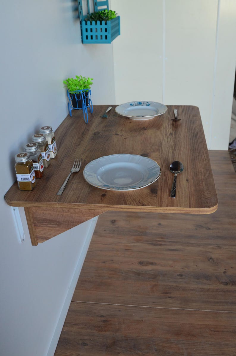 May include: A wall-mounted, wooden table with a light brown finish. Two white plates, silverware, and spice jars are arranged on the table. A small blue bicycle planter and a blue wall planter are also visible.