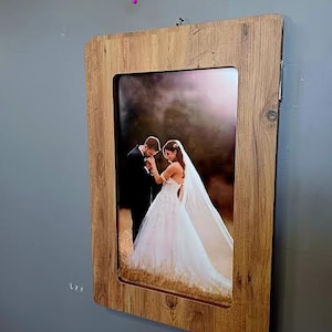 K&ouml;nnte beinhalten: Ein Holzrahmen mit einem Foto von einer Braut und einem Br&auml;utigam. Die Braut tr&auml;gt ein wei&szlig;es Hochzeitskleid und der Br&auml;utigam einen schwarzen Anzug. Das Foto ist vor einem verschwommenen Hintergrund eines Feldes aufgenommen.