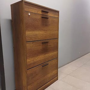 Op de afbeelding: Een bruine houten schoenenkast met vier lades en zwarte metalen handgrepen. De kast staat op een lichtbeige betegelde vloer.