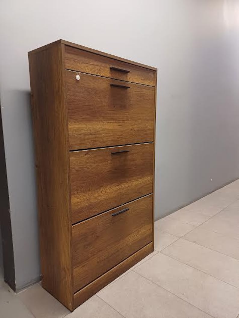 Op de afbeelding: Een bruine houten schoenenkast met vier lades en zwarte metalen handgrepen. De kast staat op een lichtbeige betegelde vloer.