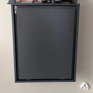 May include: A dark gray wall-mounted cabinet with a closed door. The cabinet has a simple design and is made of wood. There are toy cars and a track on top of the cabinet.