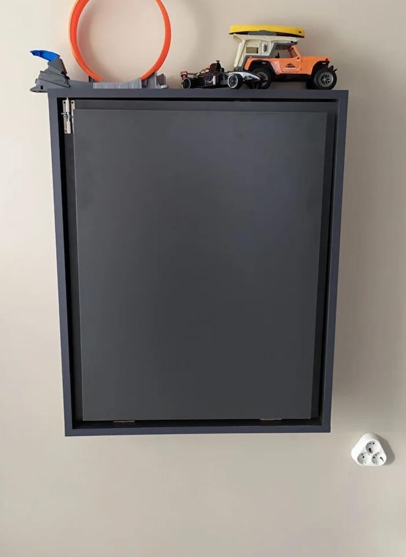 May include: A dark gray wall-mounted cabinet with a closed door. The cabinet has a simple design and is made of wood. There are toy cars and a track on top of the cabinet.