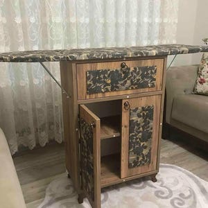 Könnte beinhalten: Ein Holz-Bügelbrett mit floralem Muster. Das Brett lässt sich herunterklappen und gibt einen Schrank mit einer Schublade und Regalen frei. Der Schrank steht auf Rollen und hat eine braune Oberfläche.