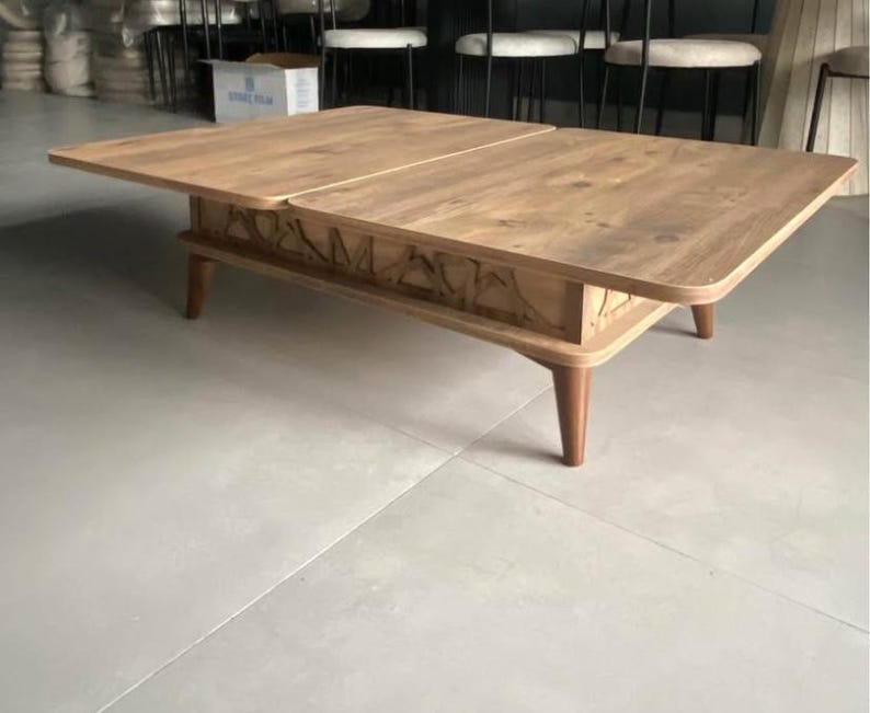 May include: A wooden coffee table with a lift-top mechanism. The table has a light brown finish and a carved design on the front. The table has four legs and is made of wood.