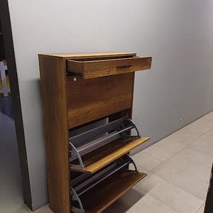 Op de afbeelding: Een houten schoenenrek met een lade en drie planken. De planken zijn open en hebben een grijs metalen frame. Het schoenenrek is bruin en heeft een houtnerf.
