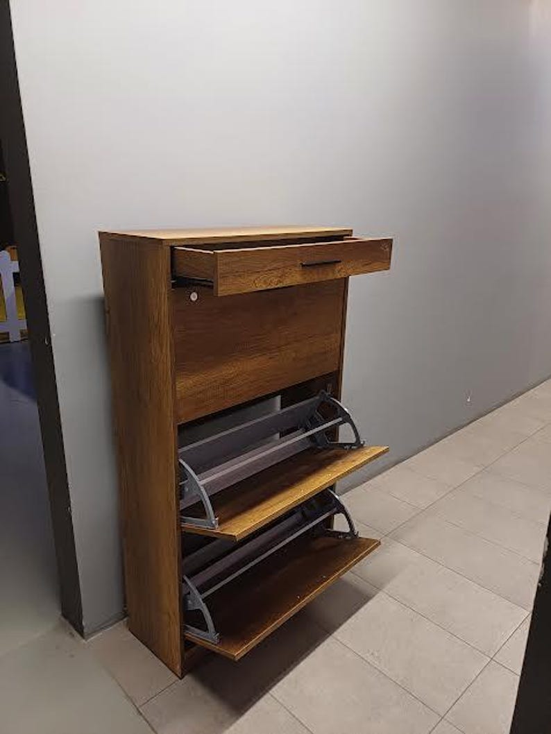 Op de afbeelding: Een houten schoenenrek met een lade en drie planken. De planken zijn open en hebben een grijs metalen frame. Het schoenenrek is bruin en heeft een houtnerf.